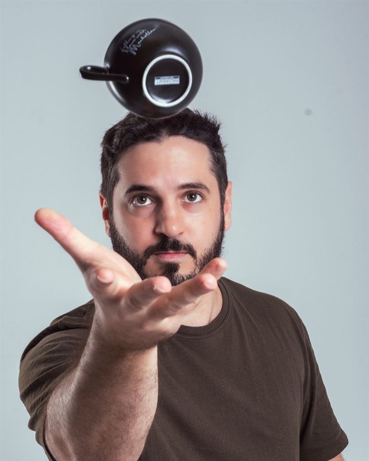 Sebastian Grau em retrato de estúdio, com uma xícara de café no ar acima da cabeça e a mão estendida para pegá-la